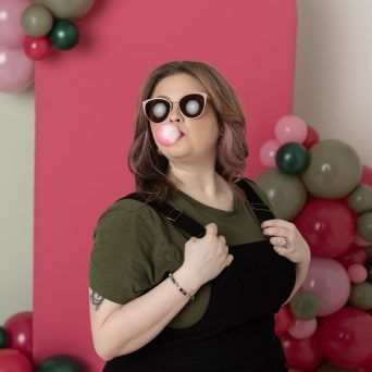 Lacey Deline in sunglasses blowing a bubblegum bubble in front of a pink balloon backdrop